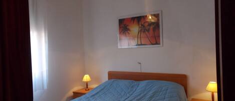 A bedroom with a bed, two bedside tables with lamps, a painting on the wall, and a window with curtains.