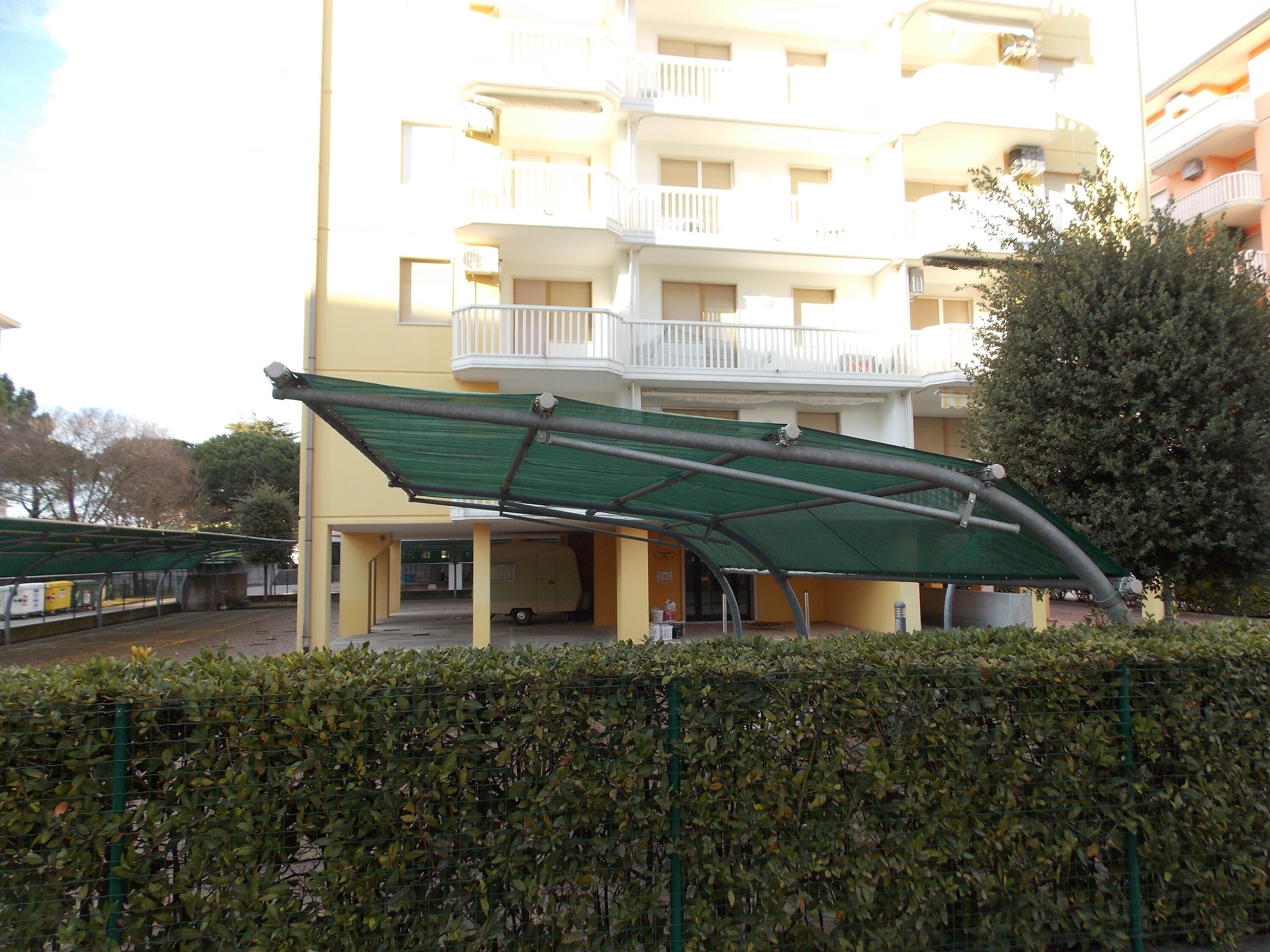 Un edificio de varios pisos con un espacio al aire libre cubierto, un balcón y un seto en primer plano.