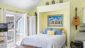 Quarto com cama, pintura de pranchas de surf, uma guitarra e vista para uma varanda.
