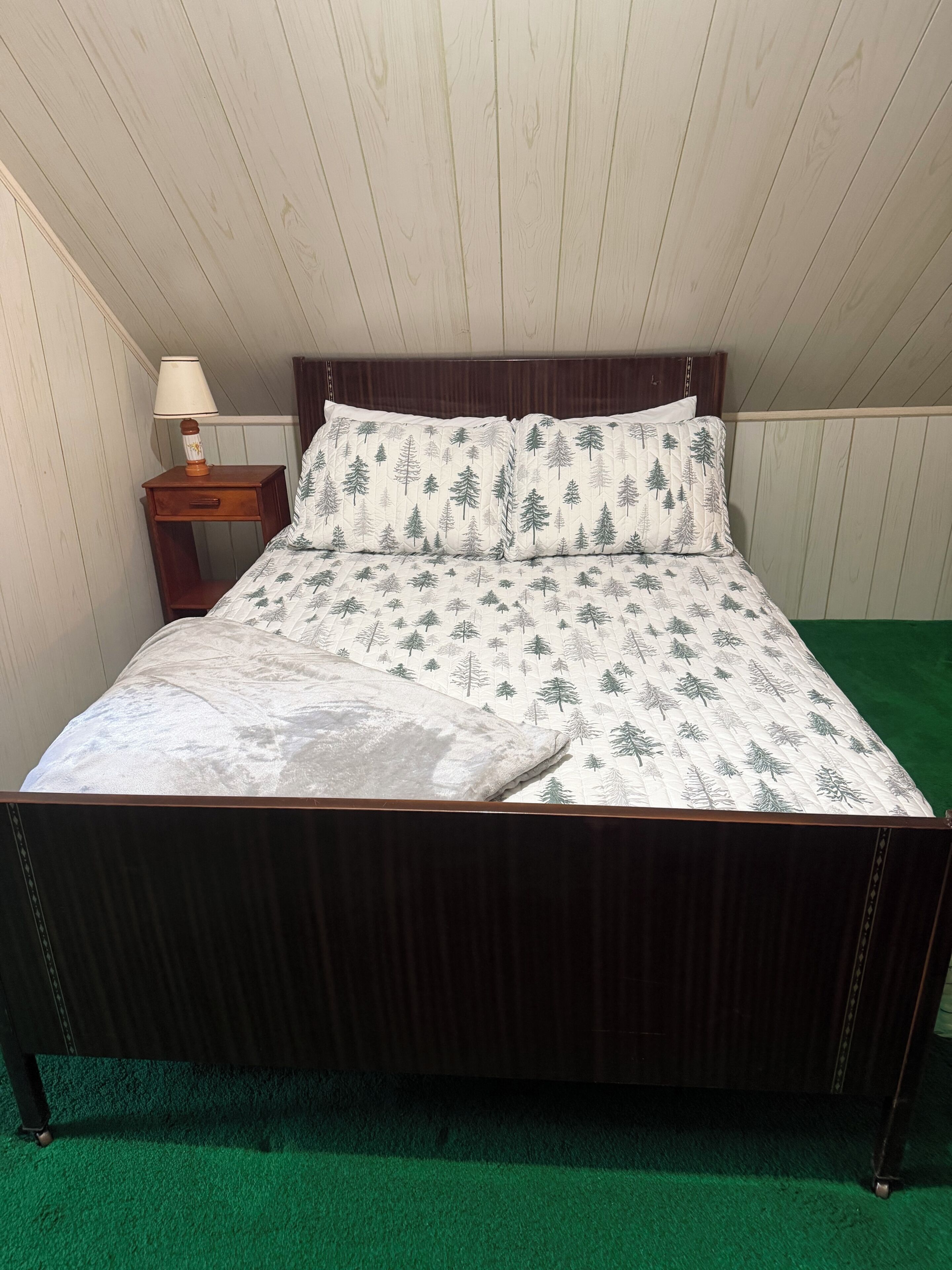 A bed with a forest pattern bedspread, a wooden nightstand with a lamp, and a green carpet.