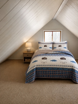 Une chambre avec un plafond en pente, un lit recouvert d’une couvre-lit à motifs, une lampe de chevet et une fenêtre.