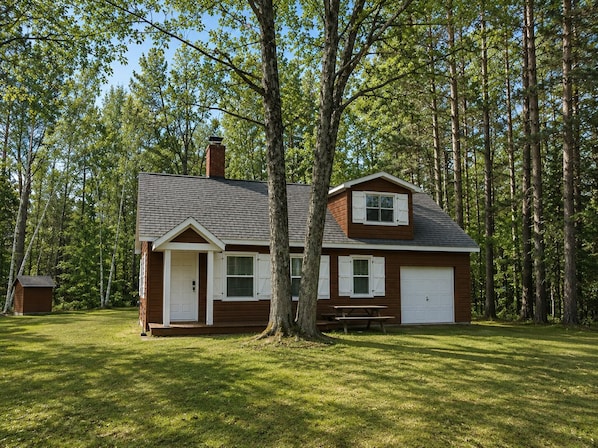 A single-story house with a front porch, a garage, and a small shed in a wooded area.