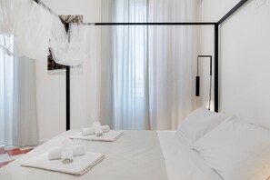 Una camera d'hotel con un letto, lenzuola bianche, struttura in metallo nero, una finestra con tende leggere e uno specchio fissato alla parete.