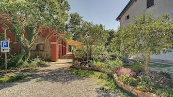 Una strada sterrata conduce a un edificio con un portico coperto, immerso in un giardino ricco di piante e fiori.
