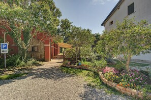 Una strada sterrata conduce a un edificio con un portico coperto, immerso in un giardino ricco di piante e fiori.
