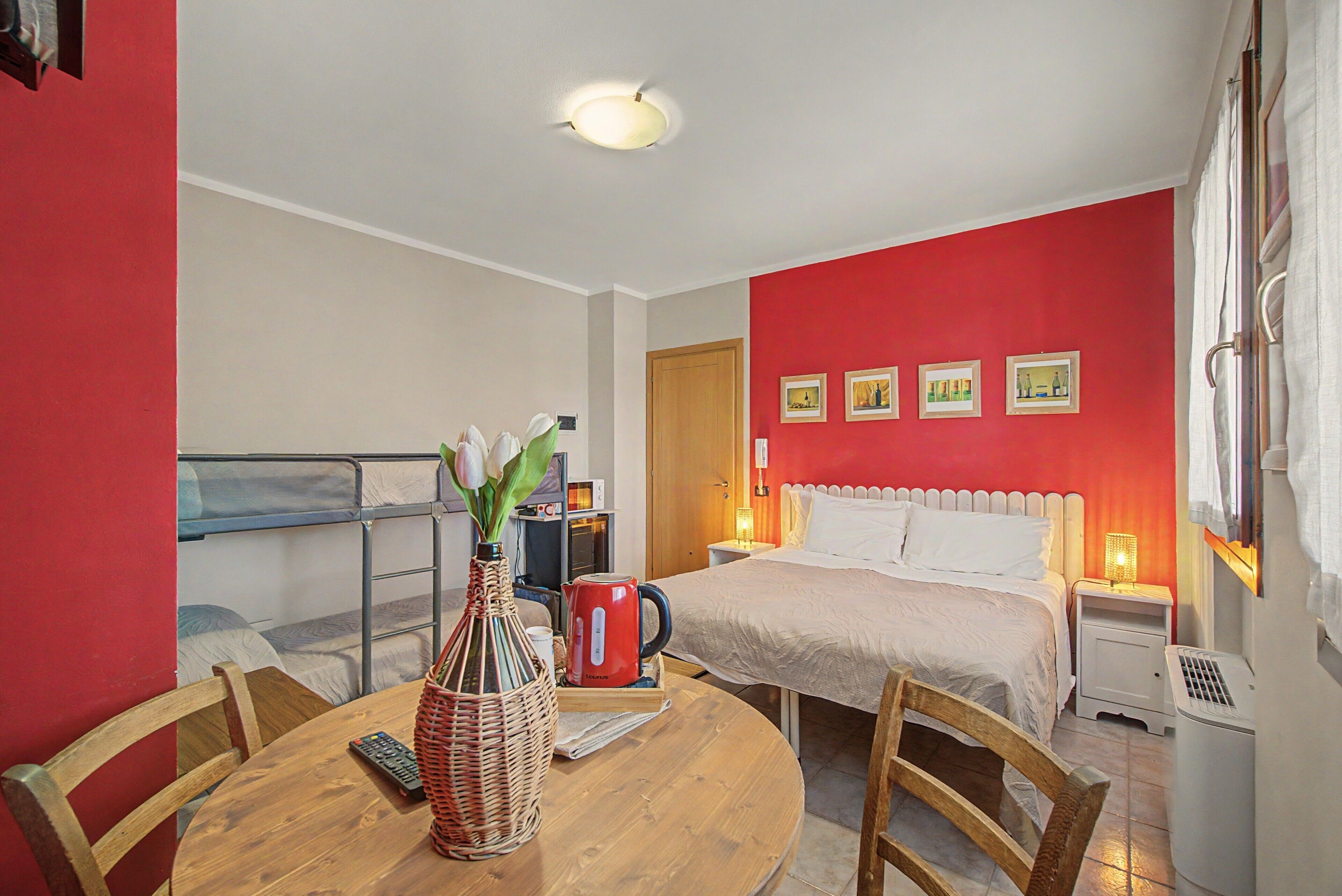 Una camera d'albergo con un letto, un tavolo con un vaso di fiori, una sedia, un muro rosso con quadri e un balcone.