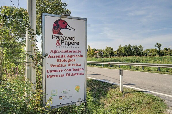 A roadside sign with text and logos, surrounded by greenery and a road.