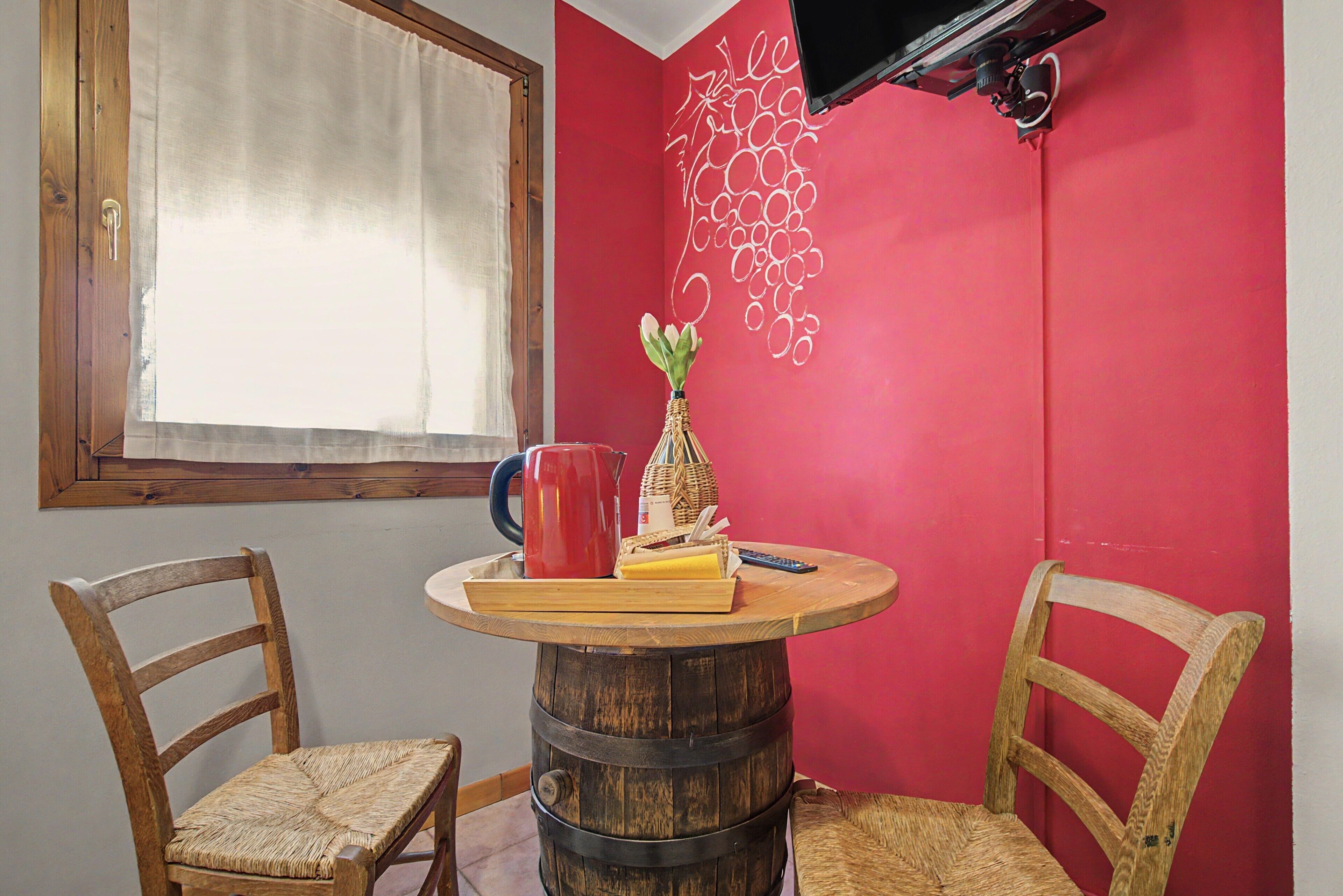 Een kamer met een ronde houten tafel, twee houten stoelen, een rode waterkoker, een vaas met bloemen, en een muur met een rood-wit ontwerp.