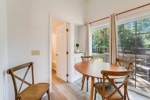 Ein Essbereich mit einem runden Tisch und Holzstühlen, einer Tür zu einem anderen Raum und einem großen Fenster mit Blick auf Grünflächen.