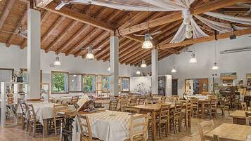 Un'ampia sala da pranzo con tavoli e sedie in legno, grandi finestre e un alto soffitto in legno con travi a vista.