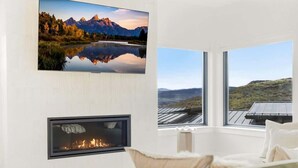 Quarto com uma televisão grande na parede, lareira e vista para uma paisagem montanhosa através de grandes janelas.