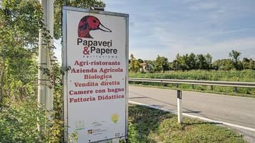 Segnaletica stradale con testo e loghi, circondata da vegetazione e una strada.
