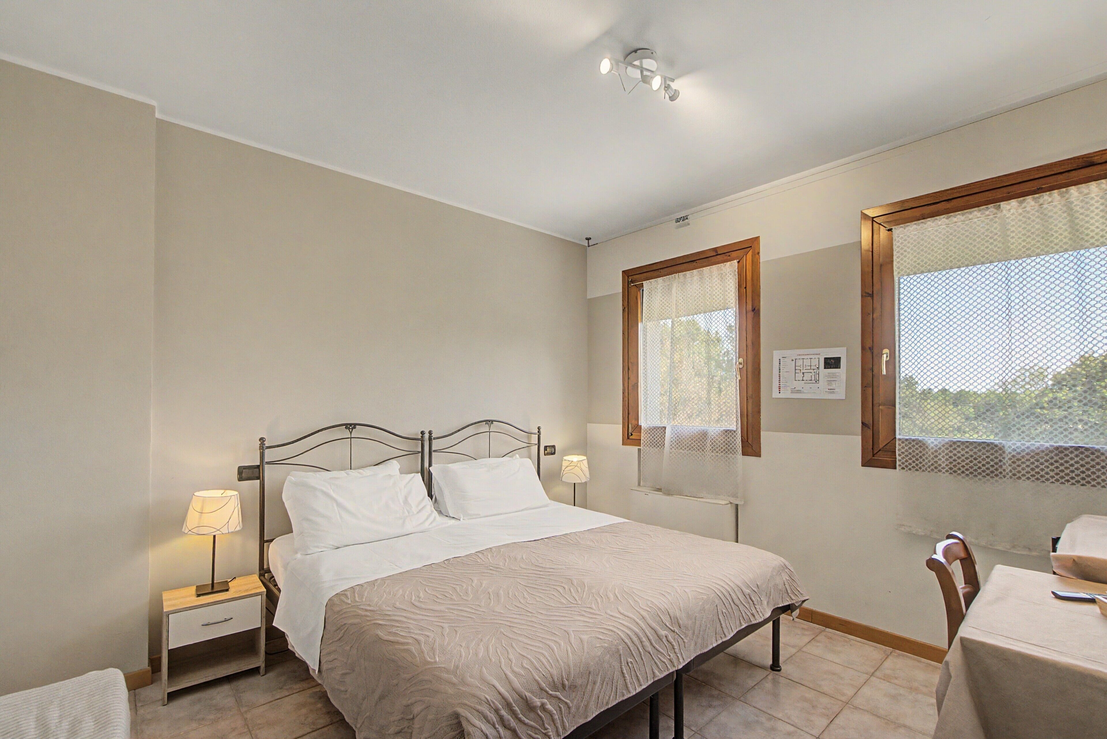Un dormitorio con cama, lámparas de noche, ventana con vistas y una mesita con silla.
