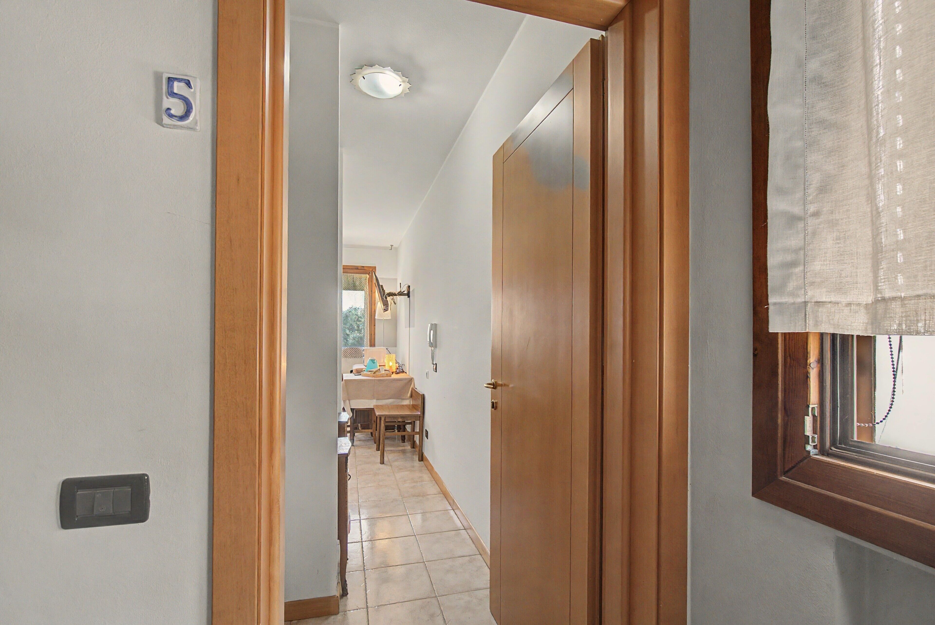 Un pasillo con puerta de madera, un cartel con el número 5, piso de baldosas y una ventana con cortina.