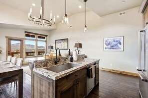 Una cocina con encimera de granito, pileta de acero inoxidable y alacenas de madera. Se observan luces colgantes, un ventanal con vistas y un refrigerador.