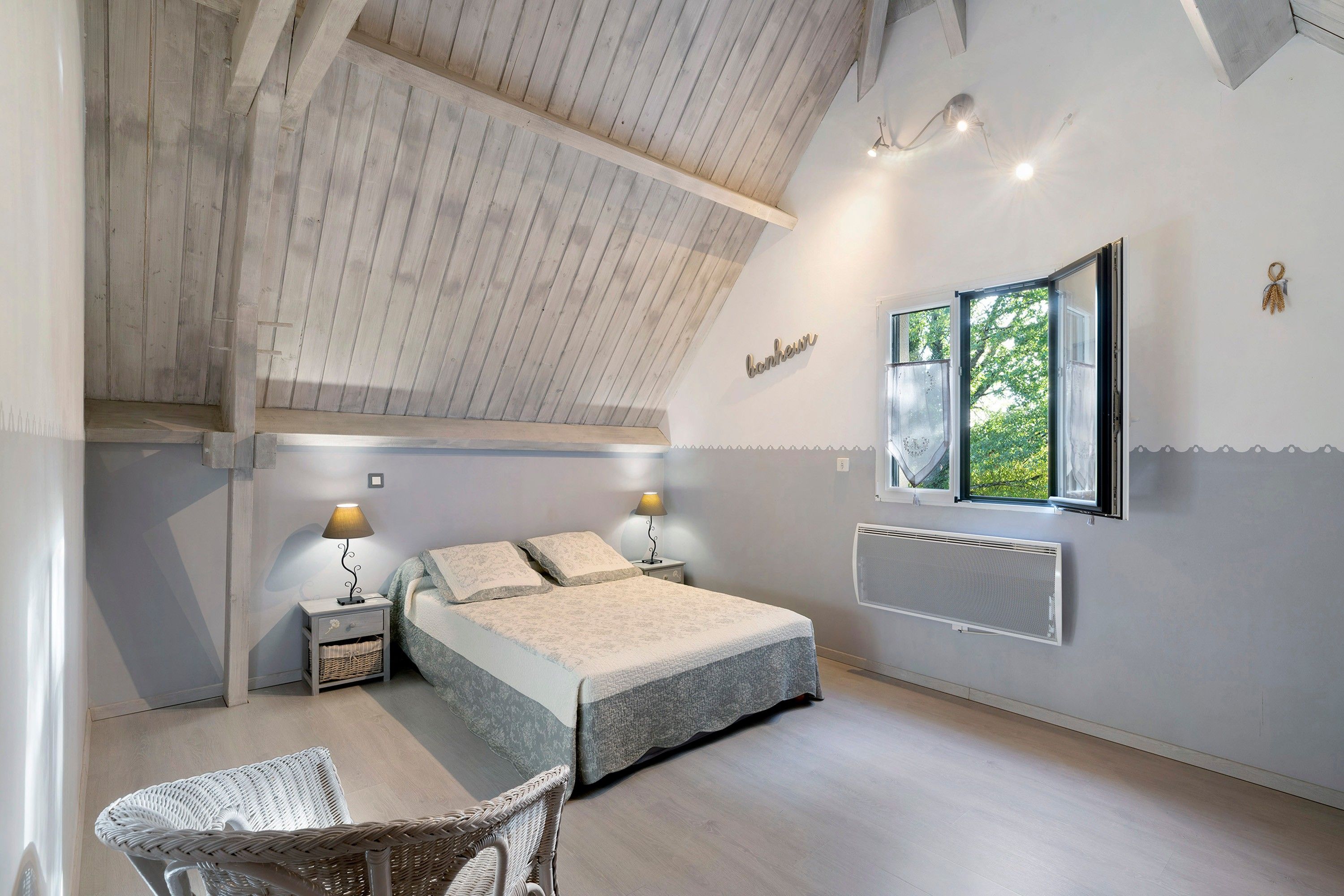A bedroom with a sloped ceiling, a bed with a patterned bedspread, two bedside lamps, a wicker chair, and a window with a view of greenery.