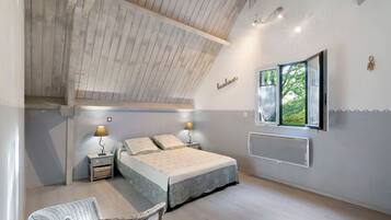 A bedroom with a sloped ceiling, a bed with a patterned bedspread, two bedside lamps, a wicker chair, and a window with a view of greenery.