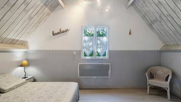 Et soveværelse med skråvæg, en seng med et mønstret sengetæppe, en senget lampe, en stol, et vindue med gardiner og en vægmonteret klimaanlæg.
