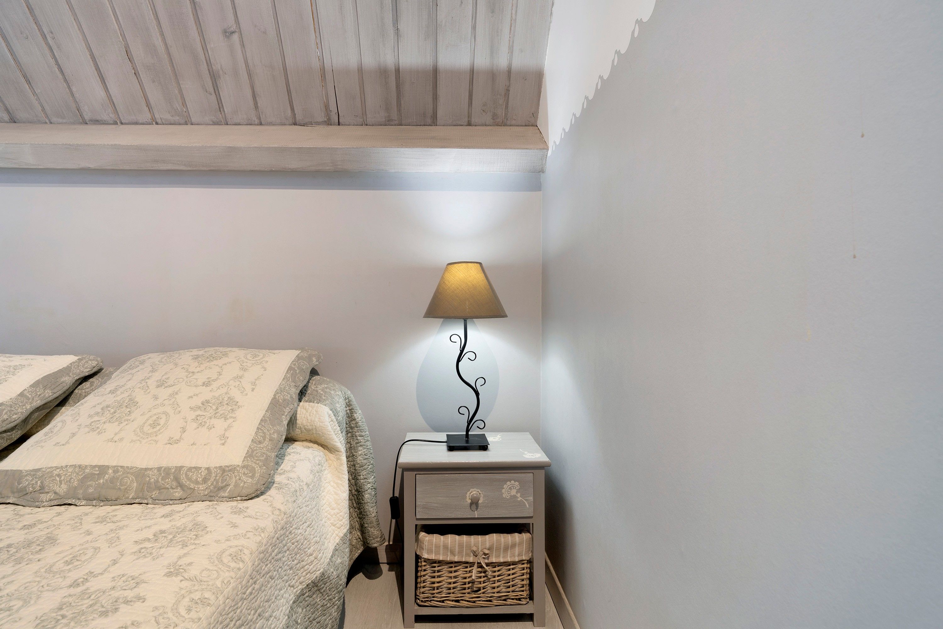 Een slaapkamer met een bed, een nachtkastje, een lamp en een klein rieten mandje.
