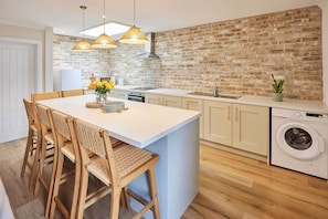 A kitchen with a brick wall, wooden chairs, a central island, and a washing machine. - Host & Stay | Puffin Cove (Seahouses)