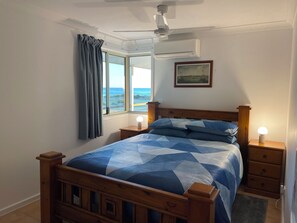 Ein Schlafzimmer mit Bett, Nachttisch, Deckenventilator, Klimaanlage, Fenster mit Vorhängen und einem gerahmten Bild an der Wand.