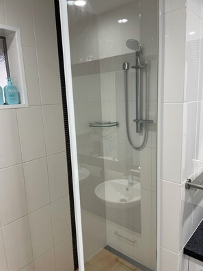 Eine Duschkabine mit einer Handbrause und einem am Wand montierten Wasserhahn, eine Glasablage mit Toilettenartikeln und eine weiße Fliesenwand.