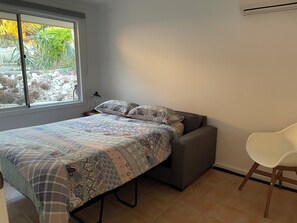 A bedroom with a bed, a chair, and a window with a view of plants and rocks.