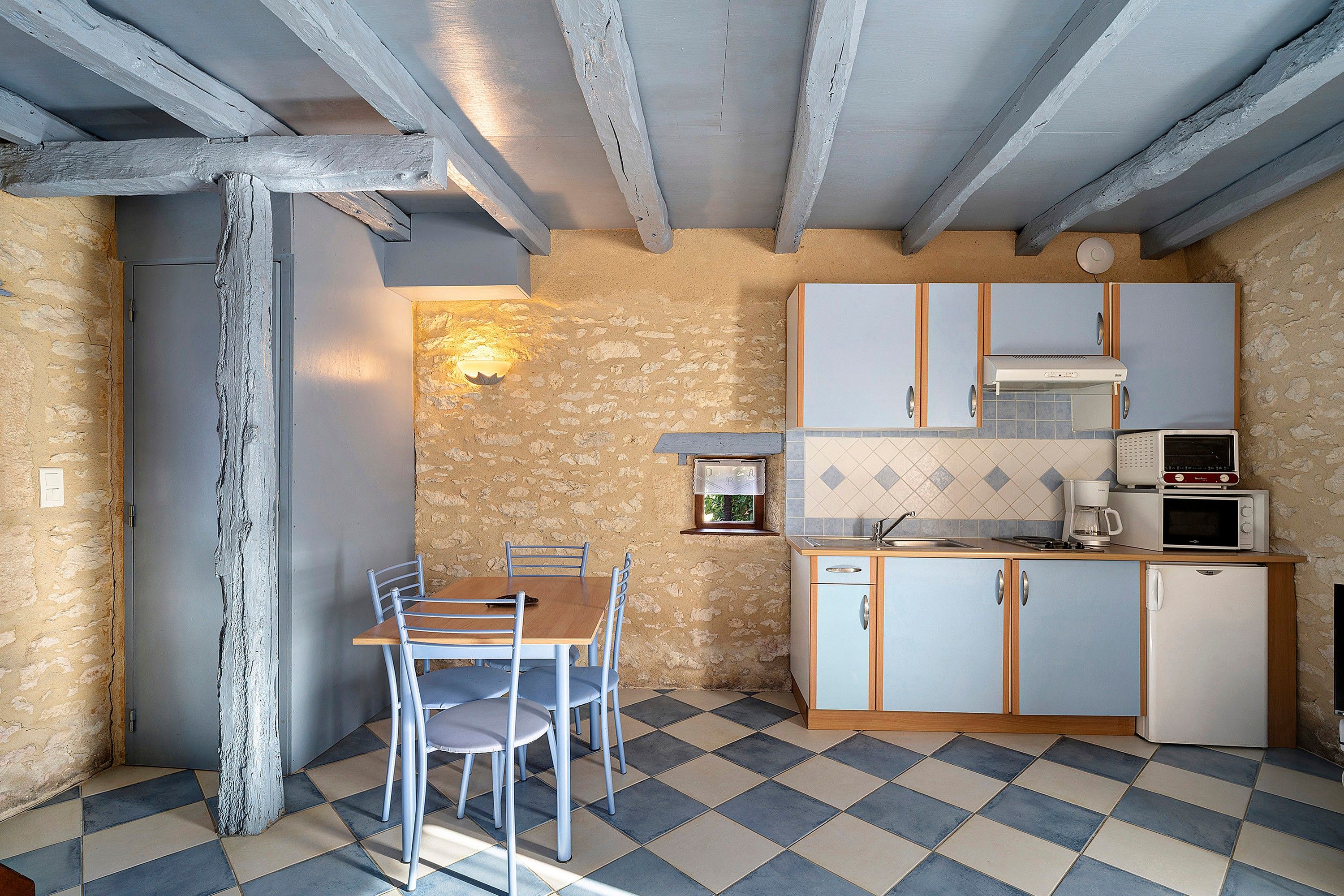 A compact kitchen with a dining area, featuring a checkered floor, stone walls, and wooden beams.