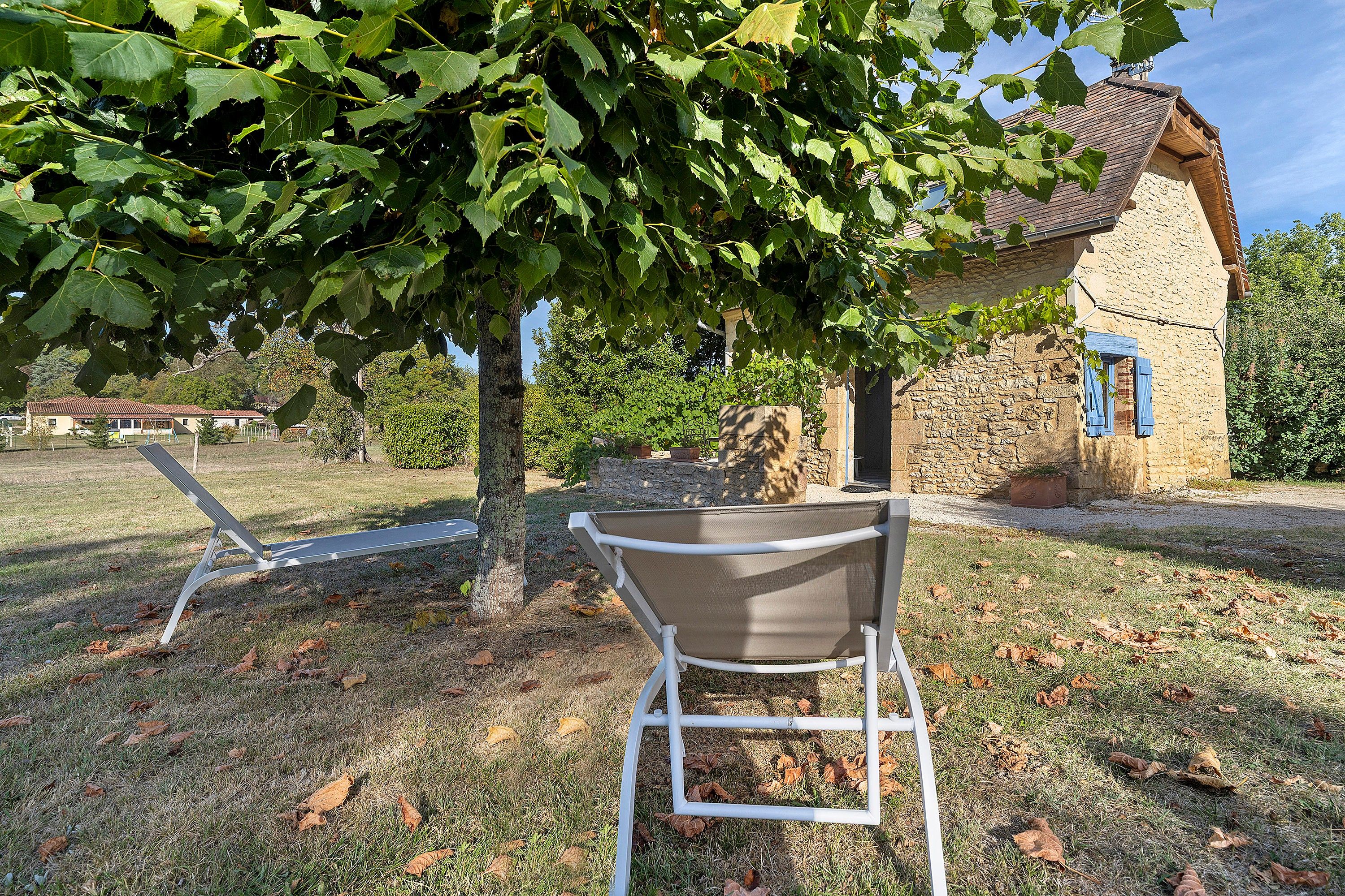 A rustic stone building with a sloped roof, a metal chair, and a tree with green leaves in a grassy area.