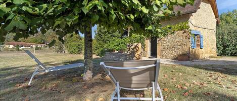 A rustic stone building with a sloped roof, a metal chair, and a tree with green leaves in a grassy area.