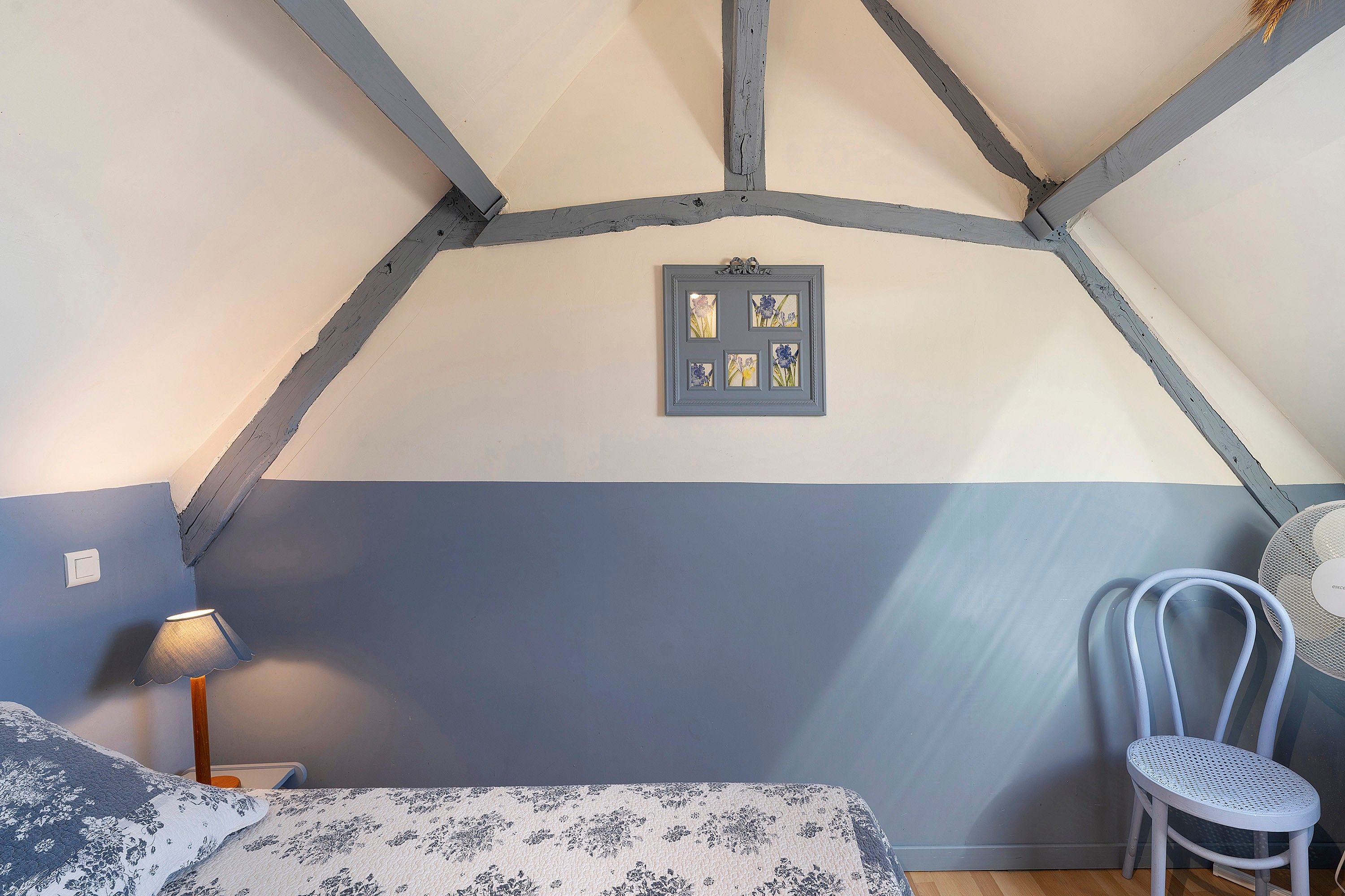 A bedroom with a sloped ceiling, a bed with a floral bedspread, a chair, and a wall-mounted picture frame.