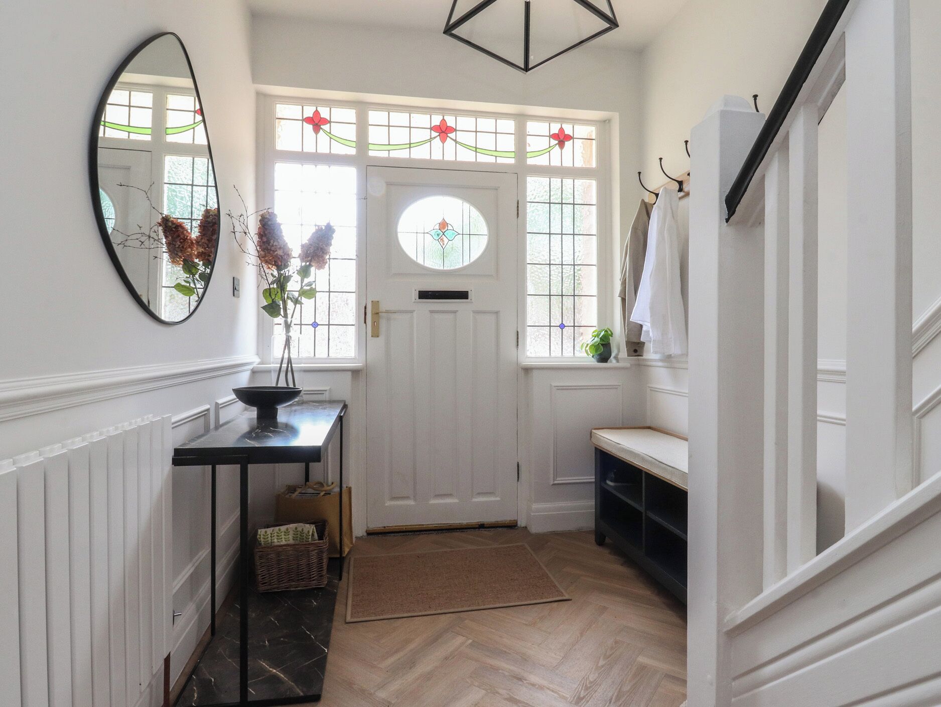 A hallway with a white door, a mirror, a vase with flowers, a small table, a basket, and a wooden floor.
