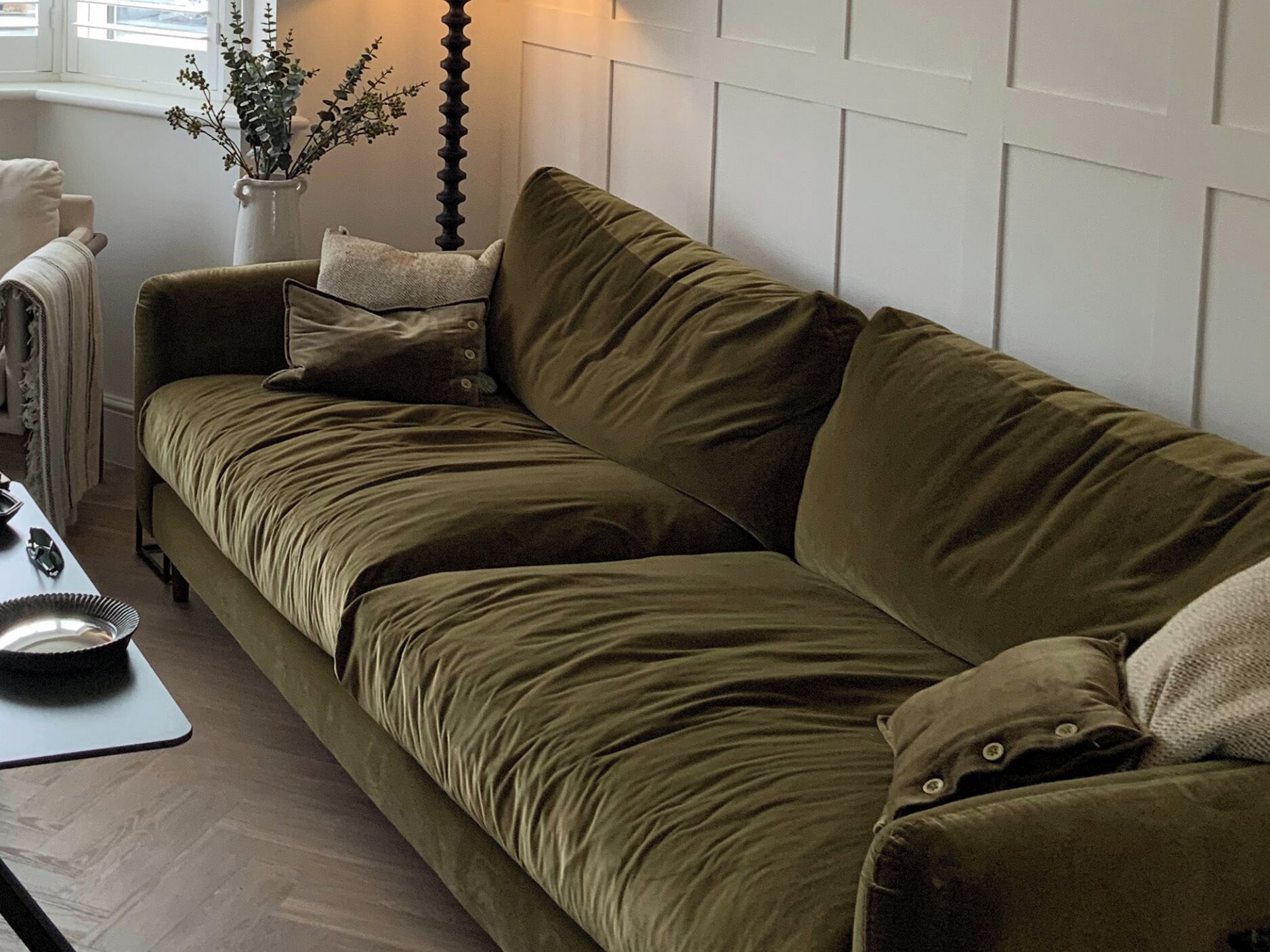 A green velvet sofa with a white cushion, a small table with a bowl and a mouse, and a white wall with a decorative vase.