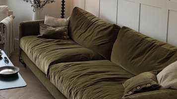 A green velvet sofa with a white cushion, a small table with a bowl and a mouse, and a white wall with a decorative vase.