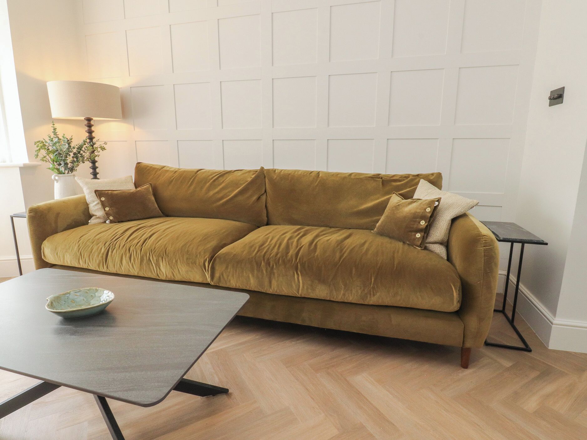 A mustard-colored sofa with cushions, a small table with a bowl, a lamp, and a plant.