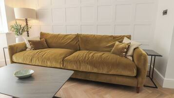 A mustard-colored sofa with cushions, a small table with a bowl, a lamp, and a plant.
