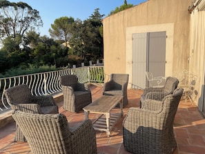 Eine Terrasse im Freien mit Korbmöbeln, einem Tisch und Blick auf Bäume und ein Gebäude.