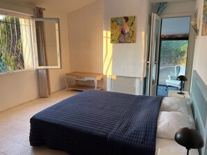 Ein Schlafzimmer mit Bett, Schreibtisch, Stuhl und Fenster mit Blick auf Grünfläche.