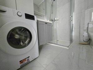 Een wasruimte met een voorlader wasmachine, een douche met glazen wand en een toilet.