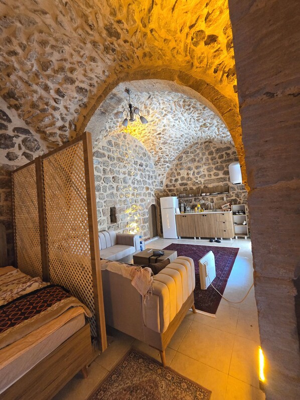 A stone-walled room with a bed, a sofa, and a kitchen area visible through an archway.
