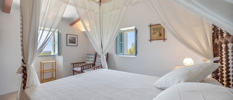 Un dormitorio con cama a dosel, cabecera de madera, mesitas de noche, una silla y ventana con vistas.