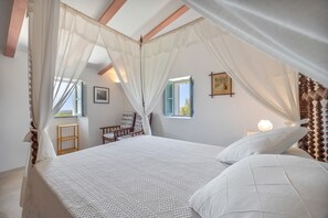 A bedroom with a canopy bed, wooden headboard, bedside tables, a chair, and a window with a view.