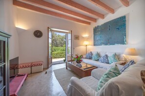 A spacious living room with a sofa, a coffee table, and a bench. There is a painting on the wall and a window providing a view of greenery outside. - Holiday Home 'Ca Na Tonina' with Sea Views and Private Pool (S'Alqueria Blanca)