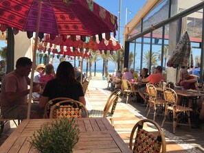 Een terras buiten met rieten stoelen en tafels, parasols en uitzicht op het strand.