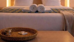 Uma cama bem arrumada com lençóis brancos, cabeceira com iluminação embutida e uma pequena mesa com um cesto de fibra.