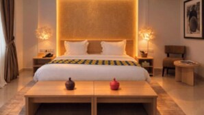 Una habitación de hotel con una cama, mesitas de noche, lámpara, una silla y una pequeña mesa con adornos.