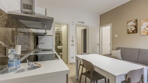A compact kitchen with a built-in stove, a dining area with a table and chairs, and a grey sofa. - Nova Suite - Apartment 200 (Calatafimi)