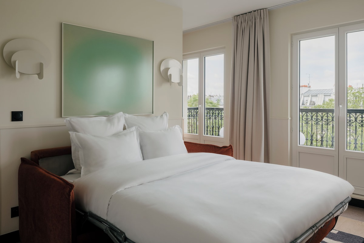 A hotel room with a large bed, a view of the city through the windows, and a green abstract painting on the wall.