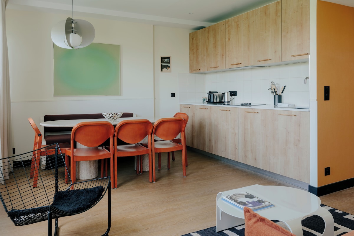 A modern kitchen with wooden cabinets, a dining area with orange chairs, and a black chair with a mesh backrest.