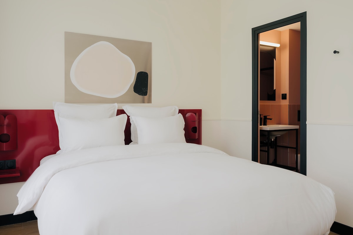 A hotel room with a bed, a red headboard, a white wall, and a door leading to a bathroom.